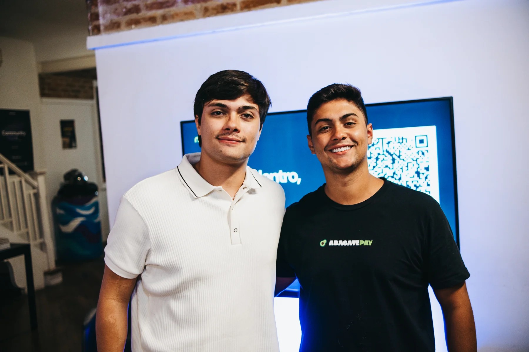 Two people pose in front of the event screen; one is wearing an AbacatePay T-shirt.