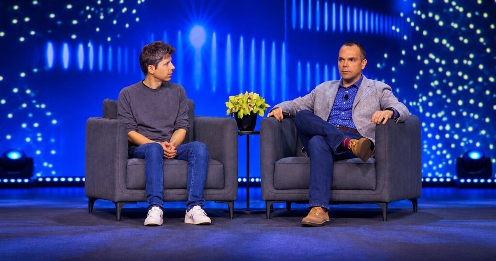 Two people speak from armchairs on stage in front of a blue screen.