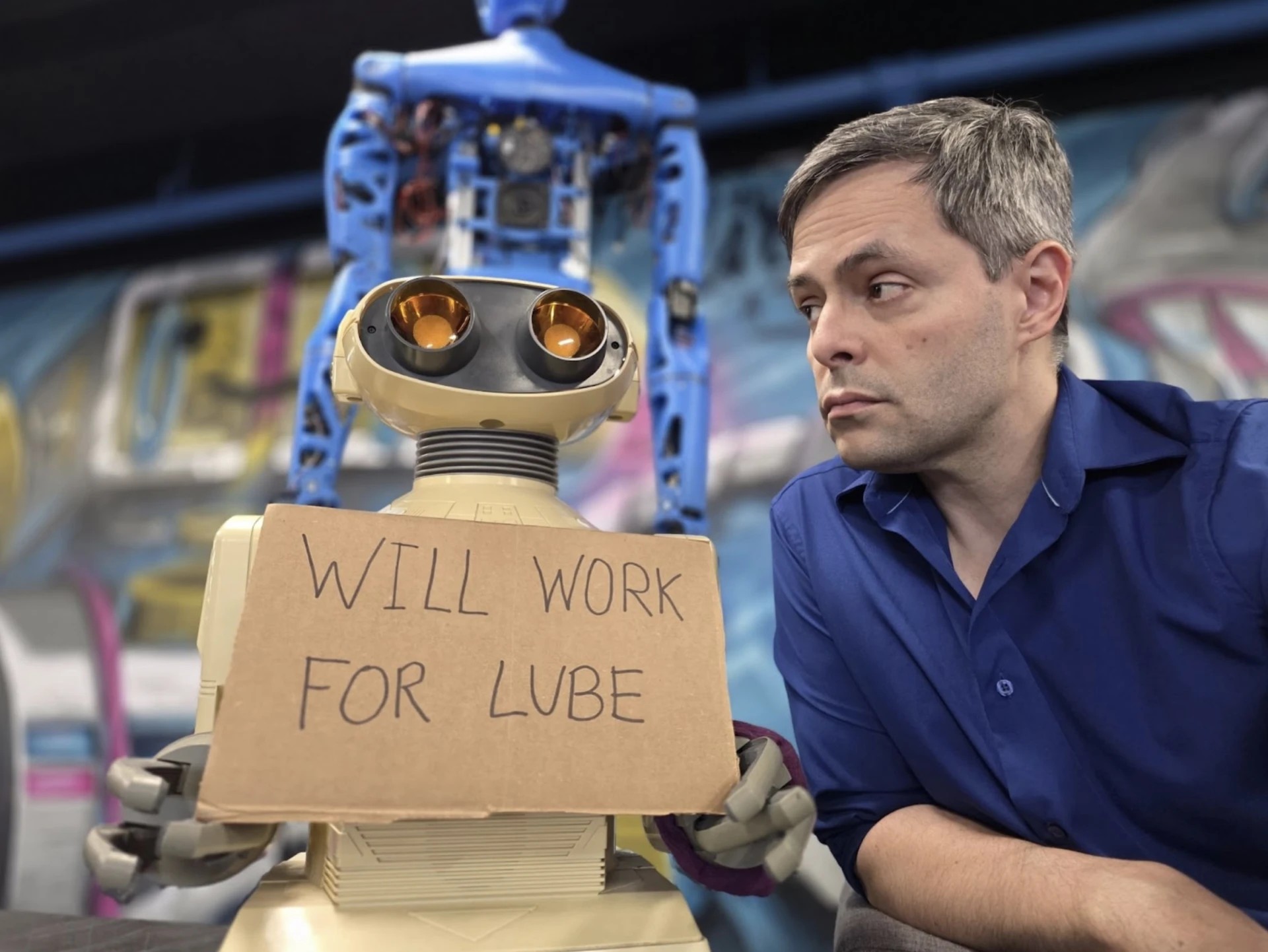 Alex Dantas ao lado de um robô que segura uma placa com a frase WILL WORK FOR LUBE.