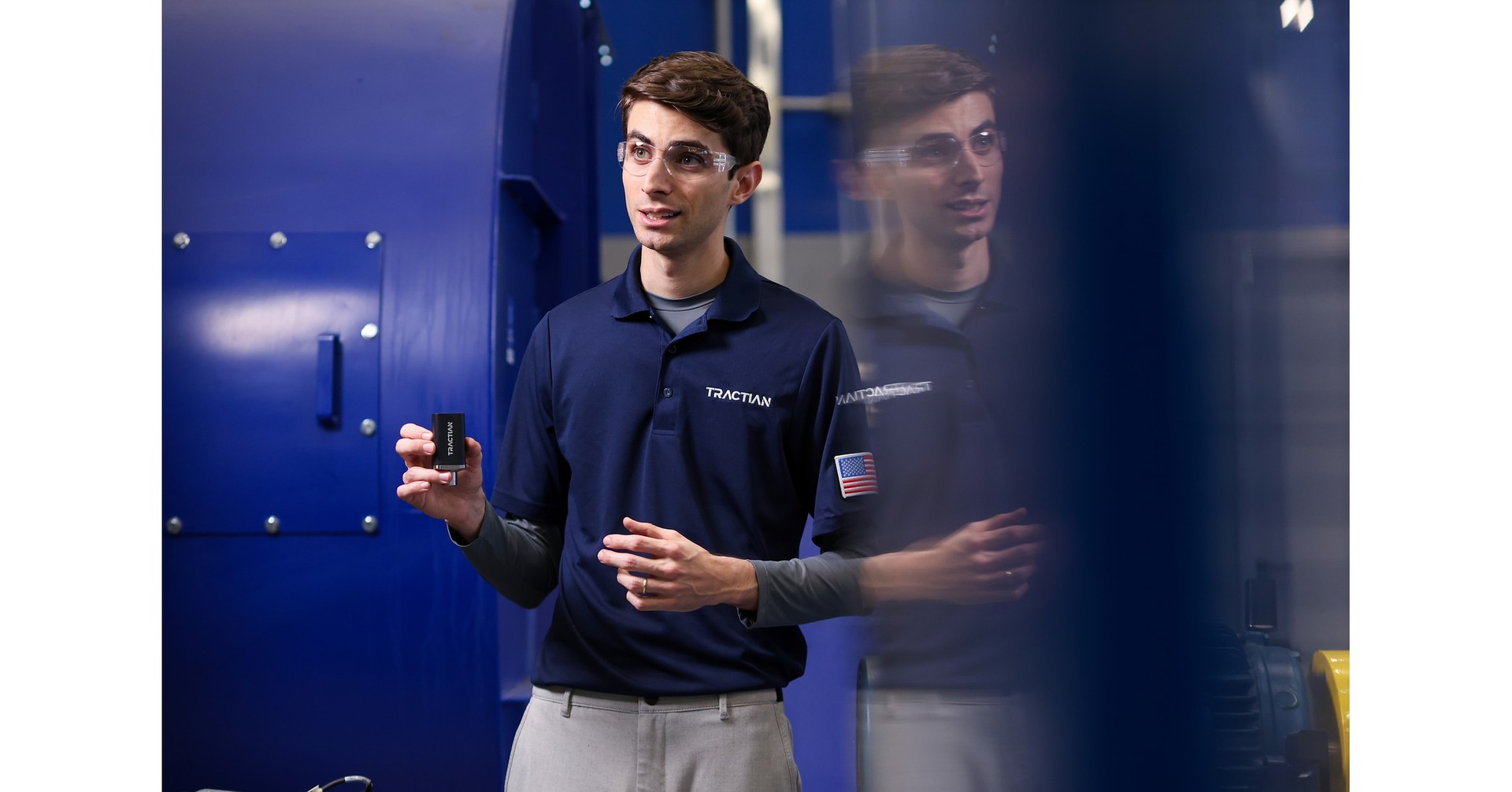 Profissional com uniforme Tractian segura um sensor diante de um equipamento industrial azul.