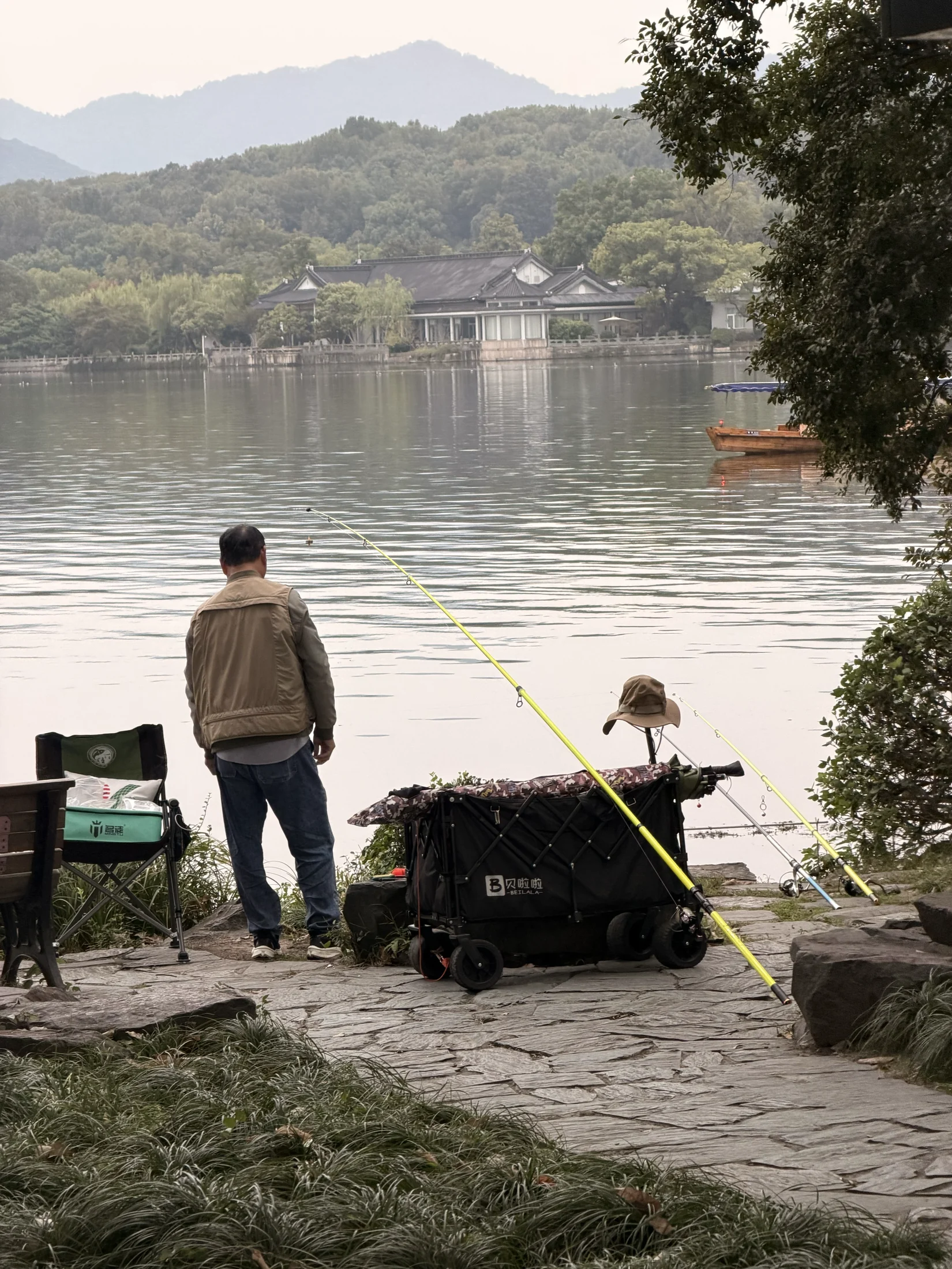 Cena de pesca à beira d’água, com construções e colinas ao fundo.