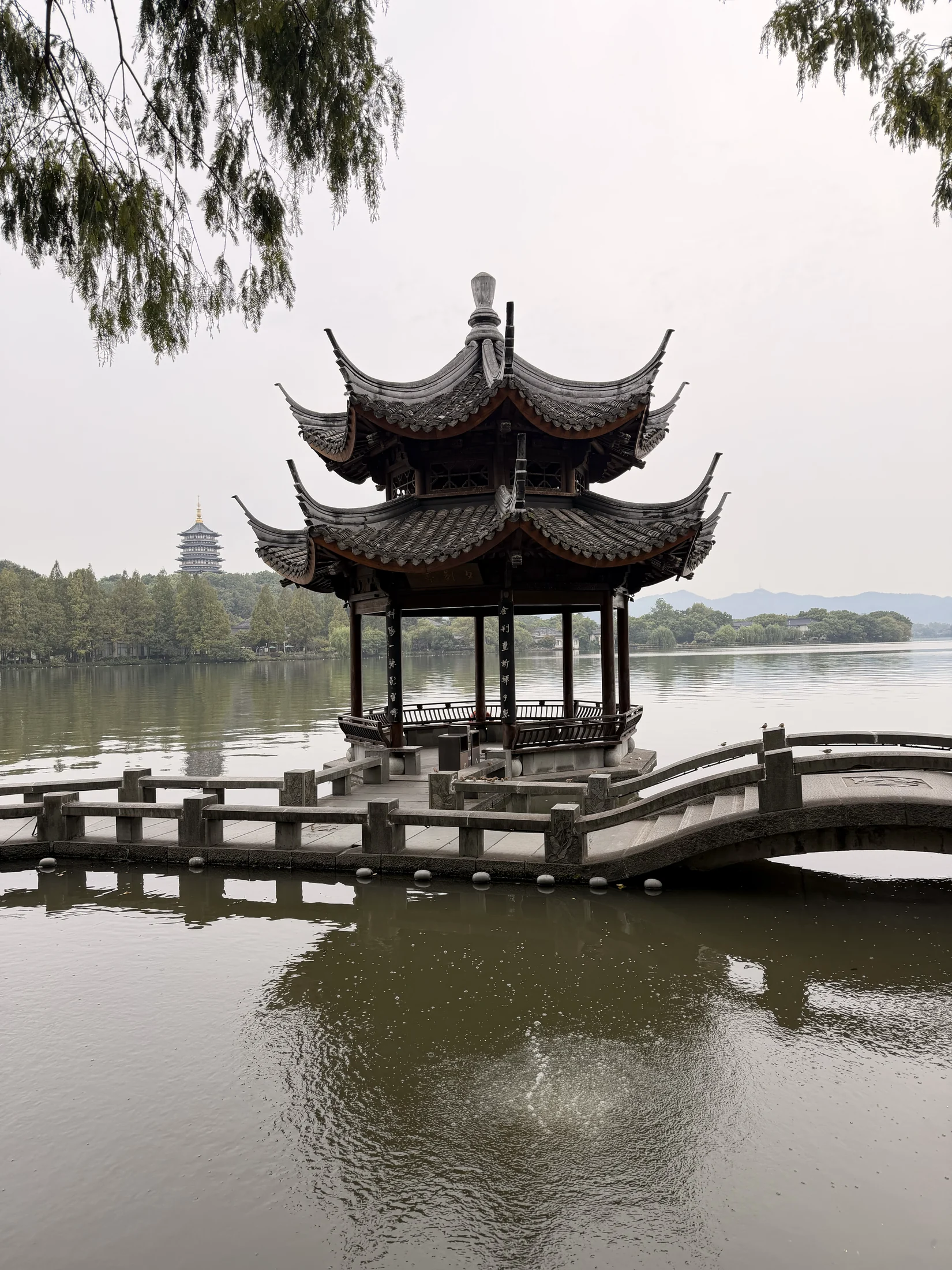 Pavilhão junto à água, com uma ponte de pedra e a paisagem de Hangzhou ao fundo.