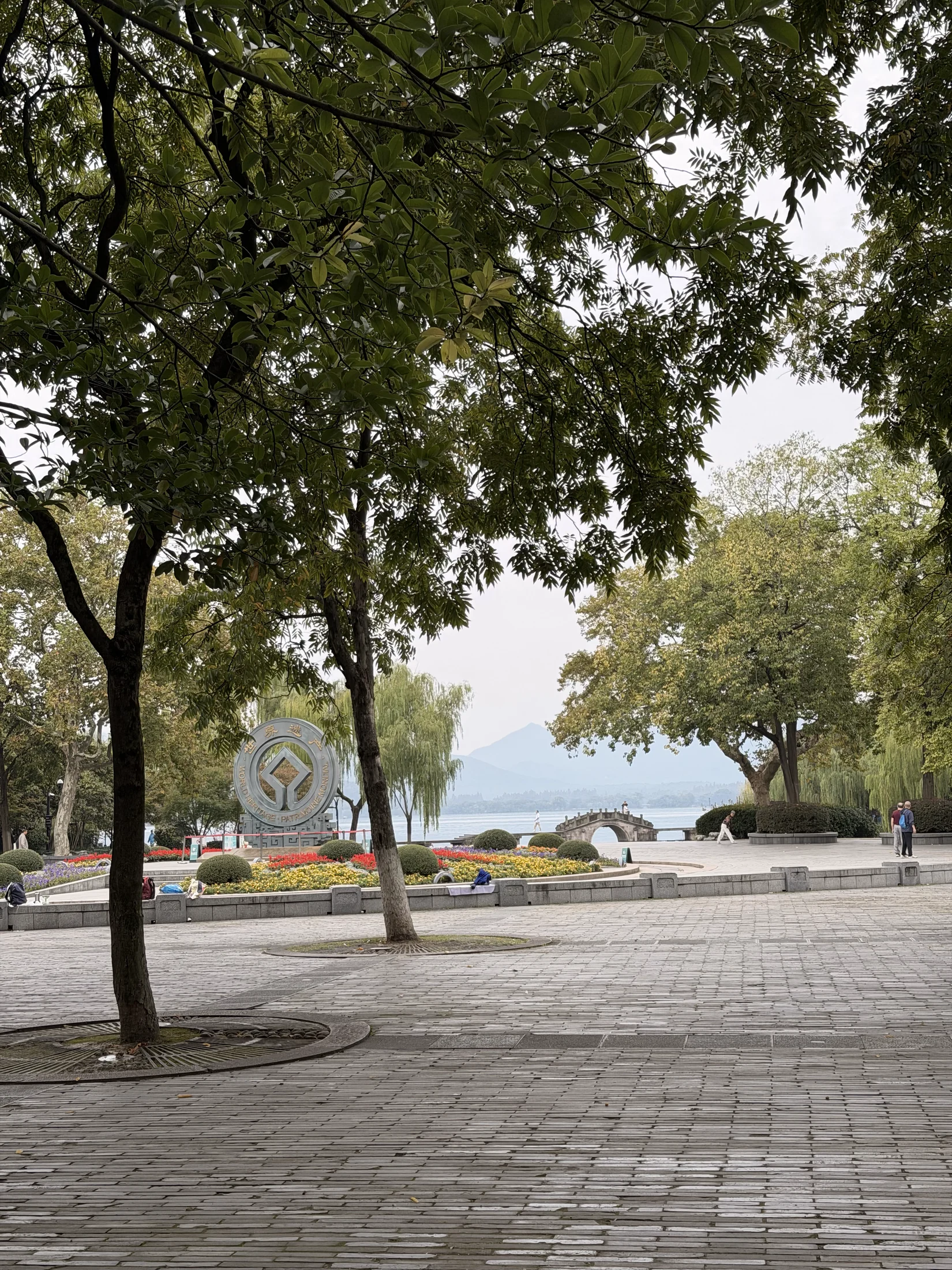 Praça arborizada com canteiros floridos e vista para a água.